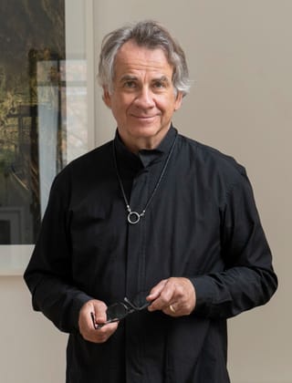 Mann in schwarzer Kleidung hält Brille, steht lächelnd vor Wandkunst.