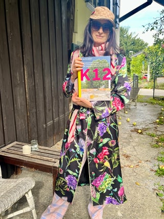 Person in floral Gewand mit Buch im Garten stehend.