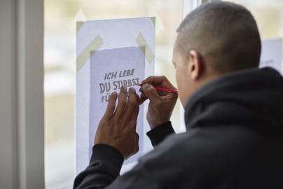 Person zeichnet Buchstaben auf Papier am Fenster.",