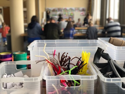 Bastelmaterial auf Tisch in Raum mit Menschen im Hintergrund.