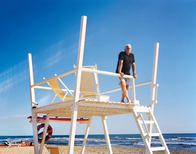 Massimo Vitali steht auf Rettungsschwimmerstuhl am Strand mit Meerblick.