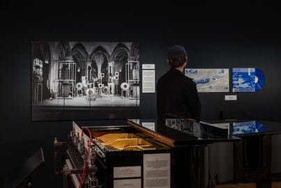 Person betrachtet Ausstellung mit Klavier und Fotos auf dunklem Hintergrund.