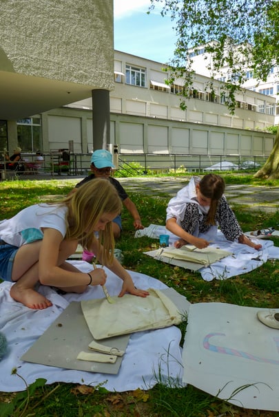 Kinder drucken draussen auf weissen Tüchern unter Bäumen.",