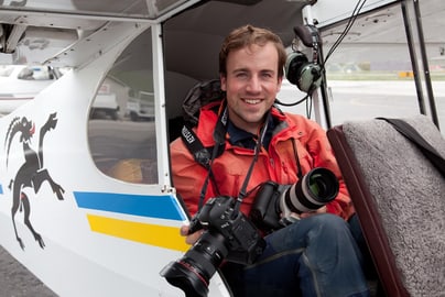 Alessandro Della Bella mit Kamera in Helikopter sitzend, lächelnd in die Kamera.