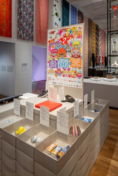Museumsausstellung mit buntem Poster und Kunstobjekten auf Regalen.