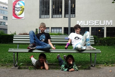 Vier Kinder sitzen auf und unter einer Bank vor einem Museum.