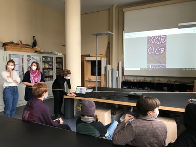 Personen mit Masken in einem Klassenraum, Präsentation läuft.",