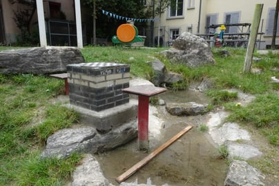 Kinderspielplatz mit Steinen und Wasserlauf in diesem eine Sitzgelegenheit aus Metall steht.