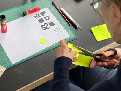 Person schneidet gelbes Papier auf Schreibtisch mit Bastelmaterialien.