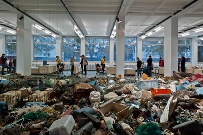 Personen betrachten in einem hell erleuchteten Raum haufenweise Kunstwerke auf dem Boden.