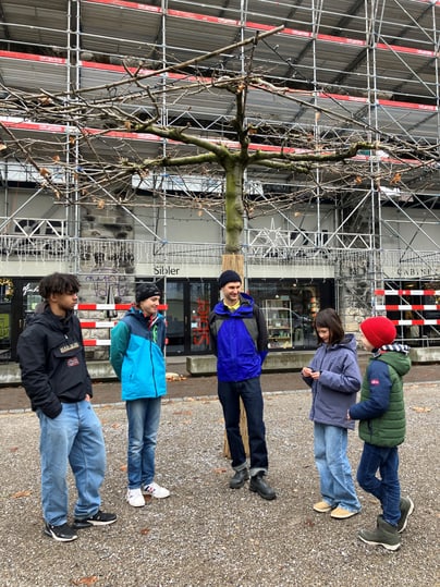 Gruppe von fünf Jugendlichen und Kindern unter einem Baum vor einem Gebäude mit Gerüst.