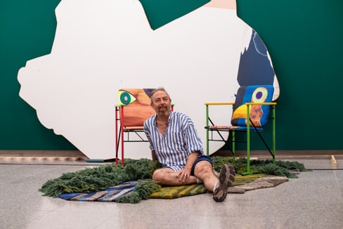 Person in gestreiftem Hemd sitzt auf Teppichen vor bunten Stühlen und Kunstwerk.