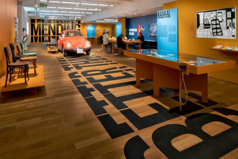 Museumsausstellung mit orangefarbenem Auto und Text auf Boden.