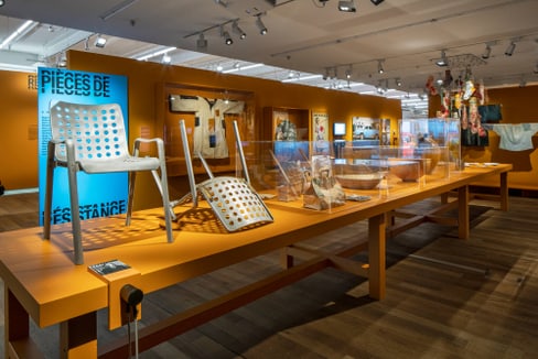Museumsausstellung mit Designstühlen und Kunstwerken auf einem langen Tisch.