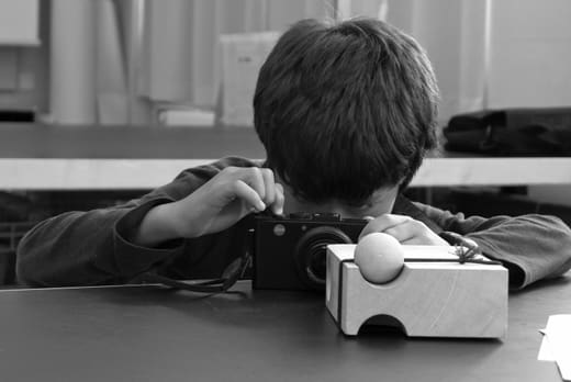 Kind fotografiert mit Kamera auf Tisch hinter Holzblock mit Ball.