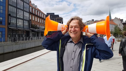 Mann hält orangefarbene Verkehrshütchen an Ohren, Strasse im Hintergrund.