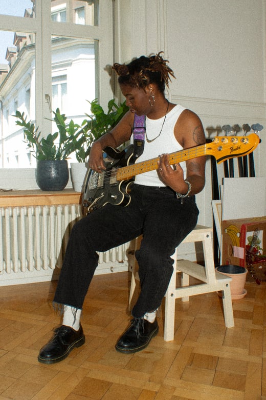 Person spielt E-Bass auf einem Stuhl sitzend in einem Zimmer mit Pflanzen im Hintergrund.