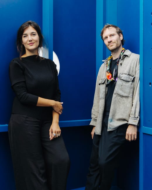 Zwei Personen stehen vor einer blauen Wand, lächelnd.