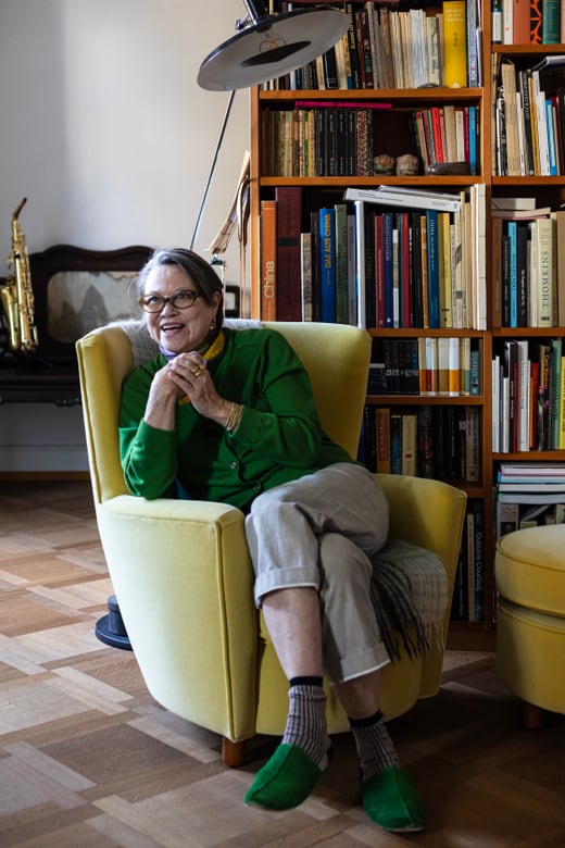 Stefi Talman sitzt auf gelbem Sessel vor Bücherregal in ihrer Wohnung. 