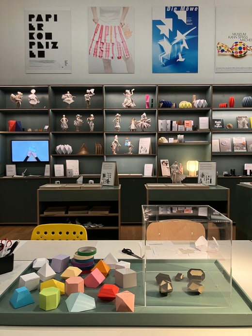 Ausstellungsraum mit Regalen und geometrischen Figuren auf dem Tisch.
